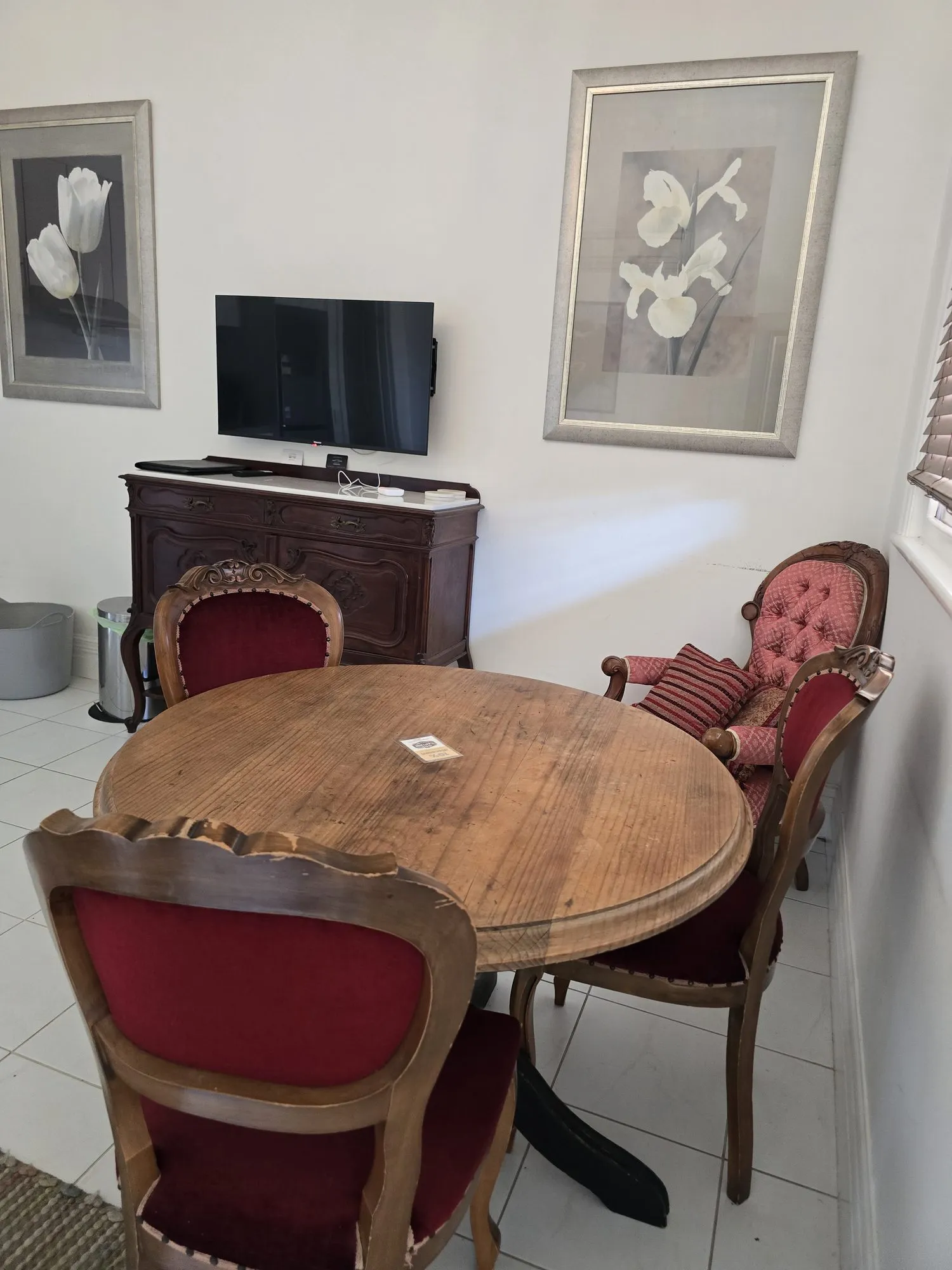 Round timber table with red velvet chairs and a buttoned armchair beside an antique sideboard