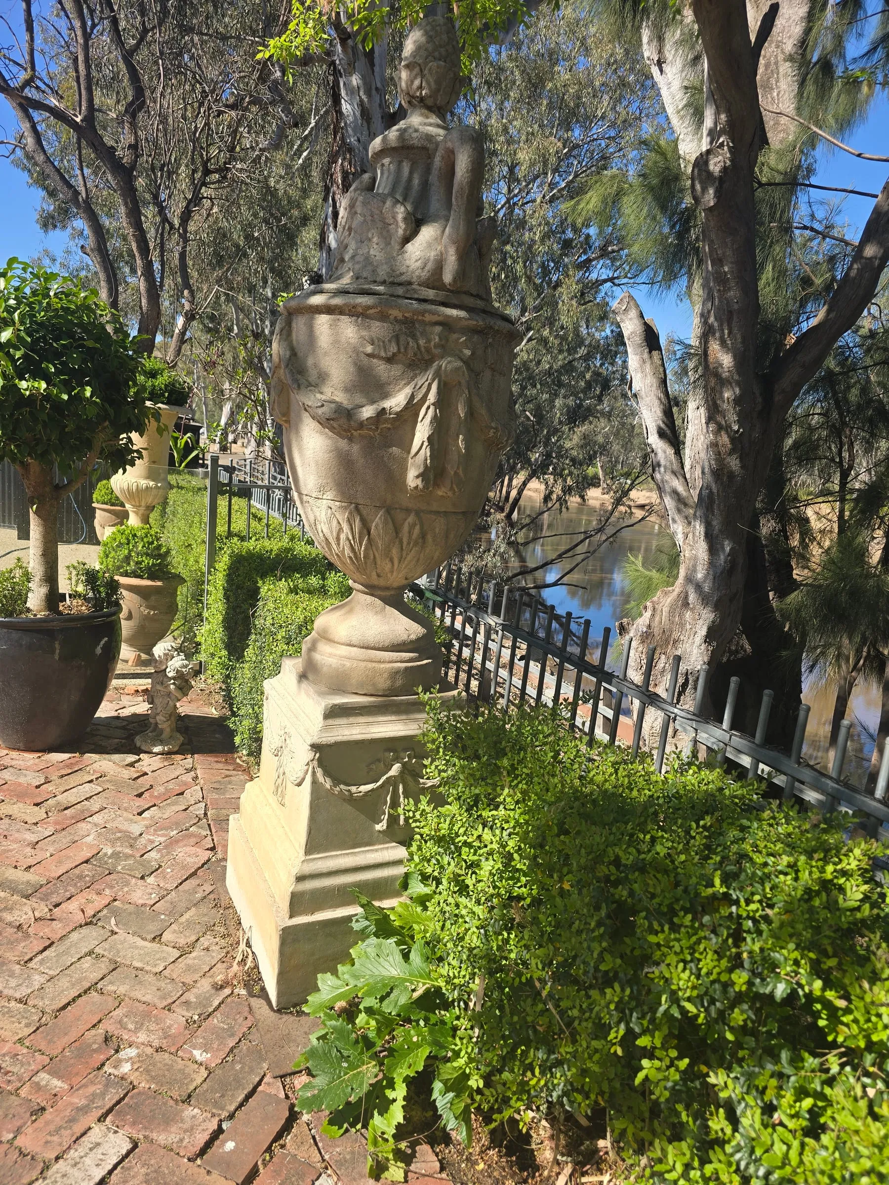 A carved stone urn on the brick garden path, the Campaspe River glinting through the gums behind an iron fence